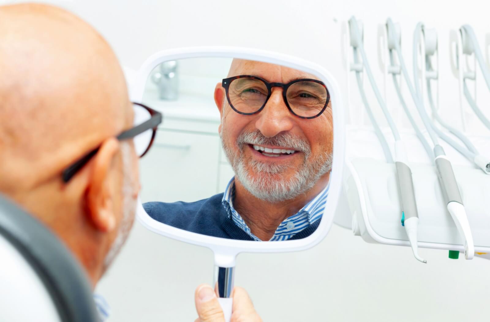 A person with grey hair and glasses smiling into a mirror at a dental clinic, showcasing the natural appearance of dental implants and tooth restoration.